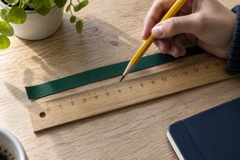A hand pointing with a yellow pencil at a wooden ruler beside a green ribbon
