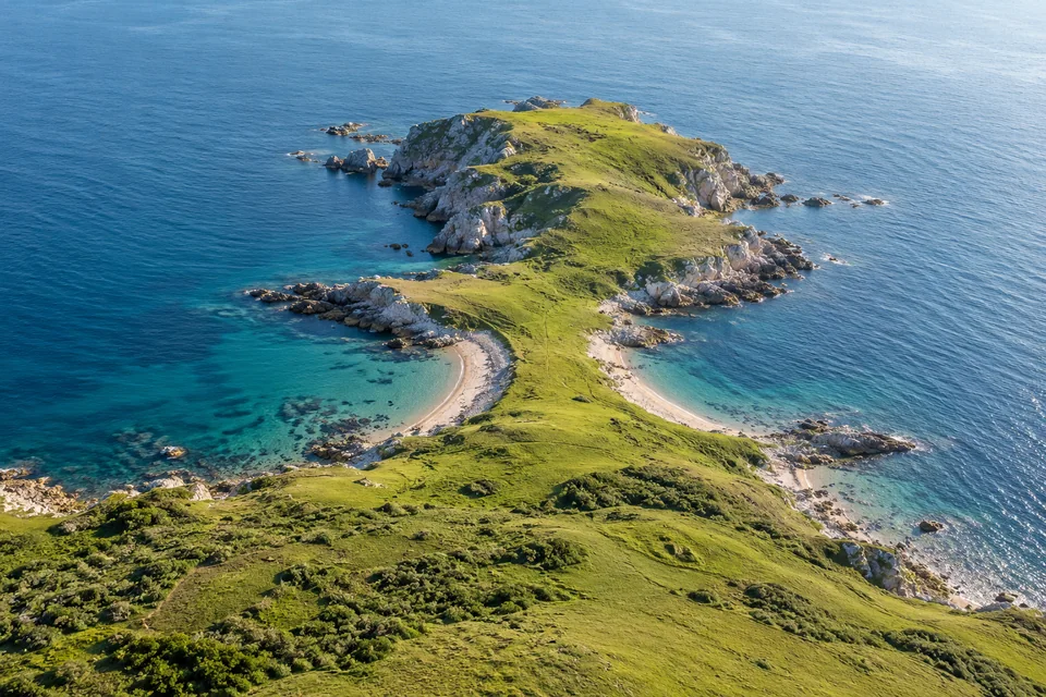 An imagined green rocky peninsula remains connected to a larger coast by a broad grassy neck, with blue sea on both sides