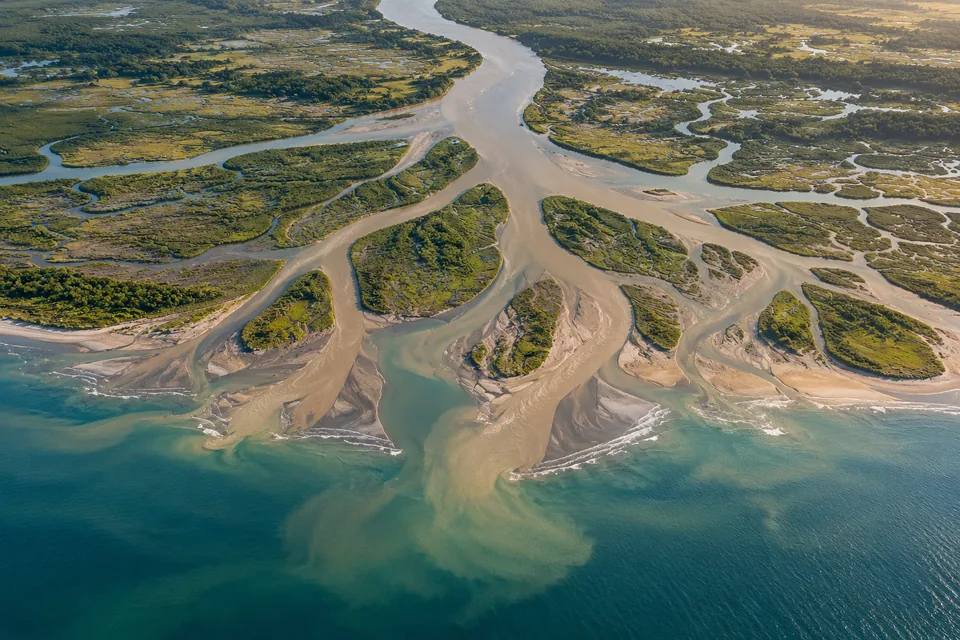 A broad muddy river divides into smaller channels around green marsh islands before meeting the sea