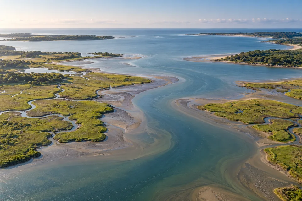 A broad tidal river channel winds between green salt marshes and mud banks into a sheltered coastal bay