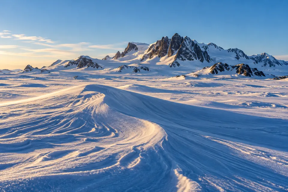 Wind-sculpted Antarctic snow ripples and blue shadows stretch towards a dark rocky mountain ridge