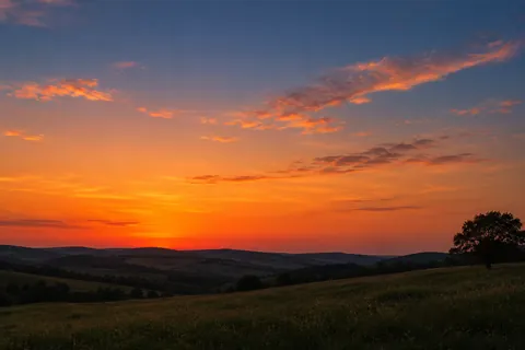 An orange and red evening sky fading into blue above quiet hills and a dark tree